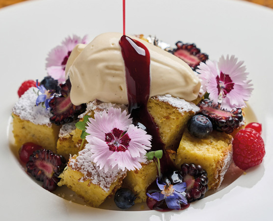 Ein geschnittenes Dessert aus saftigem Kuchenstücken, garniert mit einer Kugel Eiscreme und einem dunklen Fruchtsoß. Bunte essbare Blumen und verschiedene Beeren, wie Himbeeren und Heidelbeeren, runden die Anrichtung ab.