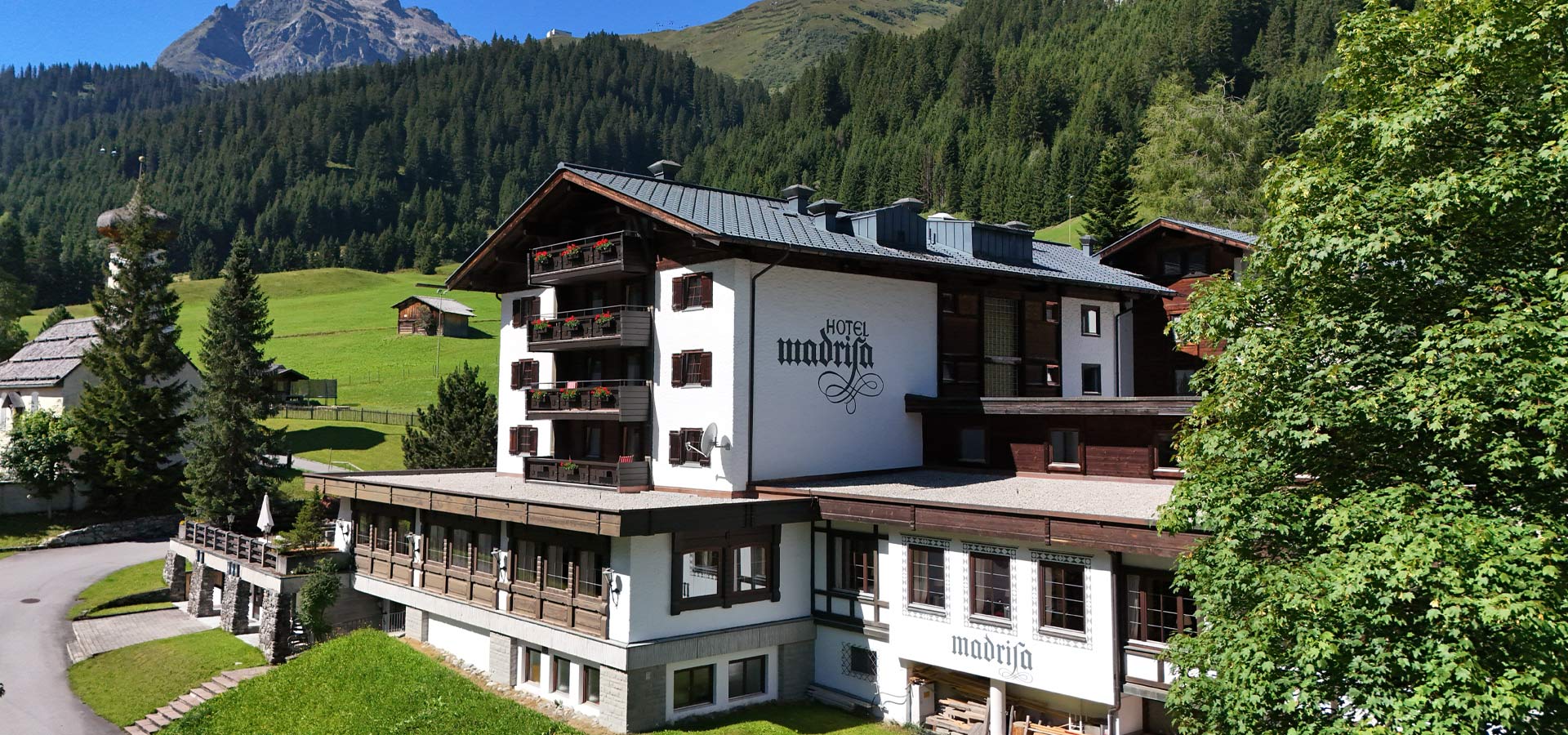 Das Bild zeigt das Hotel Madria, ein charmantes Gebäude mit mehreren Etagen und Balkonen, umgeben von üppigem Grün und Bergen im Hintergrund. Die Fassade ist in Weiß und Holz gehalten, mit dem Schriftzug „Hotel Madria“ deutlich sichtbar.