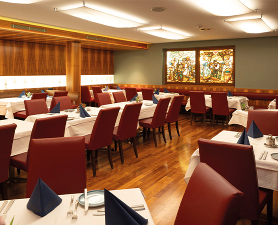 Ein elegantes Restaurant mit Holzwänden und dunklem Parkettboden. Die Tische sind mit weißen Tischdecken und blauen Servietten gedeckt. Rote Stühle umgeben die Tische. An der Wand befindet sich ein buntes Glasfenster mit figürlichen Motiven.