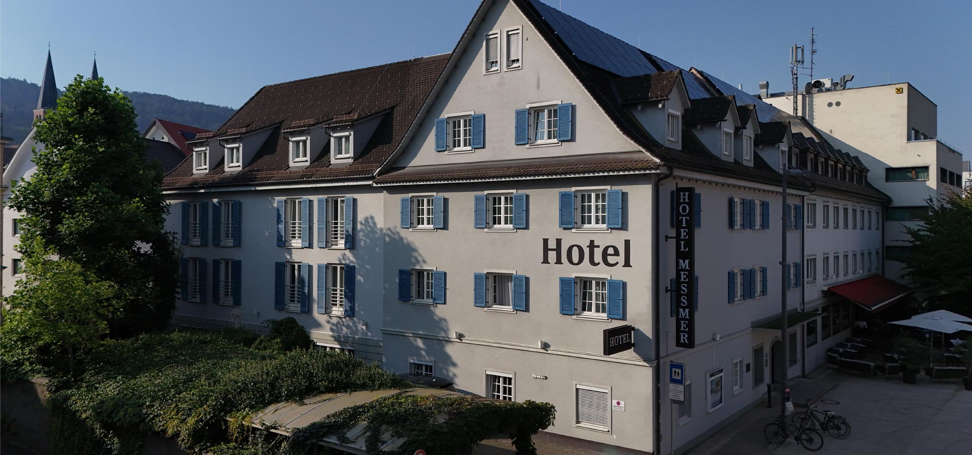 Das Bild zeigt das Hotel Messmer, ein mehrstöckiges Gebäude mit blauen Fensterläden. An der Fassade ist deutlich der Schriftzug "Hotel" zu erkennen. Vor dem Hotel befinden sich einige Bäume und eine kleine Terrasse mit Tischen und Stühlen.