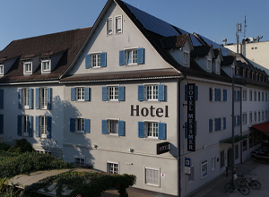 Das Bild zeigt die Fassade des Hotel Messmer mit blauen Fensterläden und einem großen Schild, auf dem "Hotel" steht. Die Architektur ist schlicht und einladend, umgeben von einer kleinen Grünfläche.