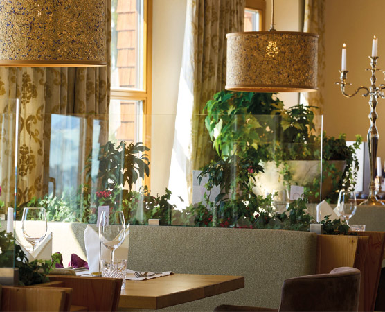 Ein stilvolles Restaurant mit eleganter Einrichtung. Auf dem Tisch stehen Gläser und Besteck. Hinter einer transparenten Trennwand sind grüne Pflanzen sichtbar. Das Licht von dekorativen Lampen schafft eine einladende Atmosphäre. Ein entspannter Ort für Gäste.