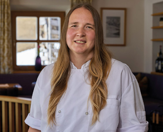 Eine lächelnde Köchin steht in der Küche, trägt eine weiße Kochjacke und hat langes, lockiges Haar. Im Hintergrund sind Fenster und einige Möbel zu sehen.