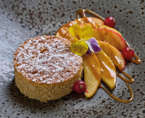 Auf dem Bild ist ein Dessert zu sehen, das aus einem runden Kuchen mit Puderzucker bestäubt besteht. Daneben liegen frisch geschnittene Äpfel und einige rote Früchte, dekoriert mit einer essbaren Blume. Das Arrangement ist auf einem strukturierten Teller angerichtet.