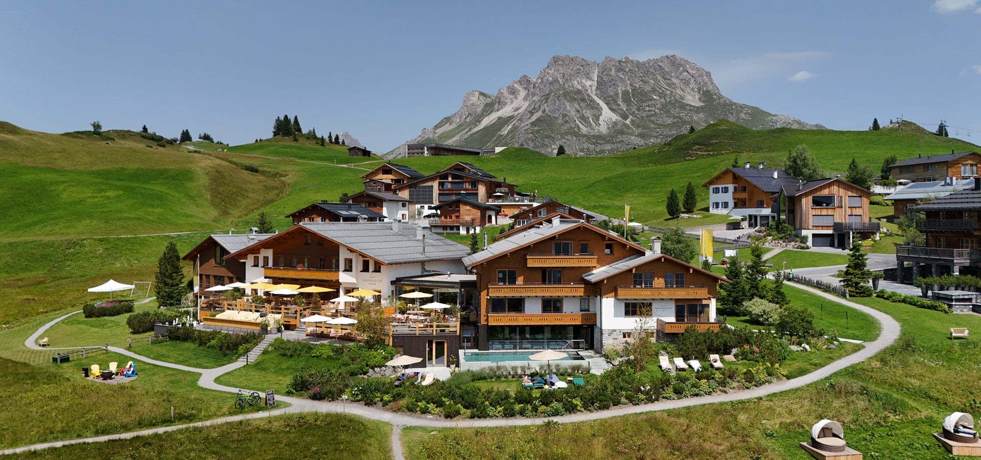 Eine malerische Alpenlandschaft mit mehreren modernen Chalets ist abgebildet. Die Gebäude sind umgeben von grünen Wiesen und Bäumen. Im Vordergrund ist eine Terrasse mit Sonnenschirmen und Liegen zu sehen, während im Hintergrund majestätische Berge aufragen.
