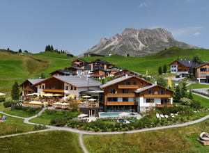 Eine malerische Hotelanlage aus mehreren Holzhäusern liegt in einer grünen Hügellandschaft. Im Hintergrund sind majestätische Berge sichtbar. Einige Terrassen sind mit Sonnenliegen und Tischen ausgestattet, um Gästen einen Blick auf die Natur zu bieten.
