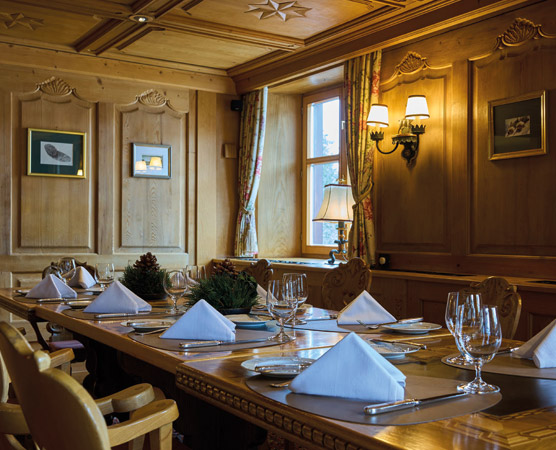 Ein eleganter Essraum mit einem langen Holztisch, gedeckt mit weißen Servietten und Gläsern. An den Wänden hängen Bilder, und große Fenster lassen Licht herein. Der Raum ist gemütlich gestaltet mit warmen Holzverkleidungen und sanften Farbnuancen.