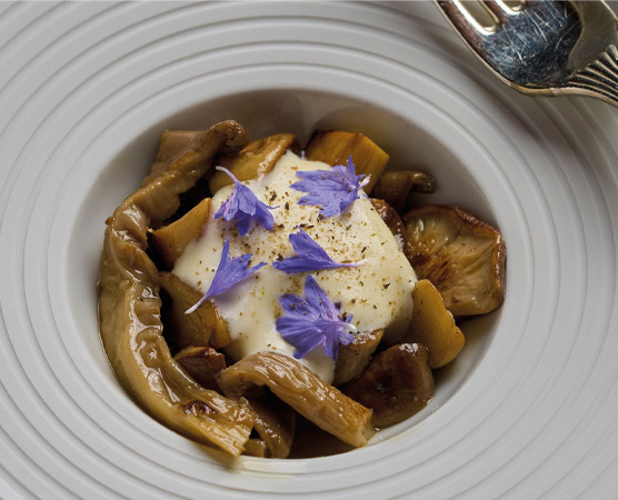 Eine elegante Schüssel enthält eine Mischung aus gebratenen Pilzen und samtiger Creme. Das Gericht ist mit essbaren Blüten dekoriert, die ihm eine farbige Note verleihen. Im Hintergrund ist ein silberner Löffel sichtbar.