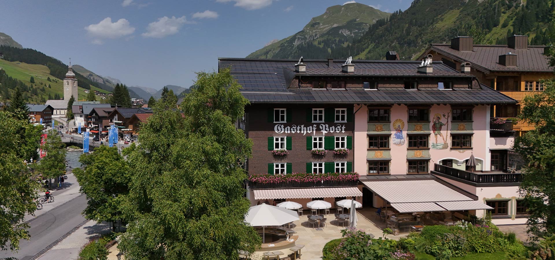 Ein traditioneller Gasthof namens "Gasthof Post" in einer malerischen Berglandschaft. Das Gebäude ist liebevoll gestaltet mit bunt blühenden Fenstern und Wandmalereien. Vor dem Gasthof befinden sich Terrassen mit Sonnenschirmen und Tischen, umgeben von Bäumen und einer schönen Umgebung.