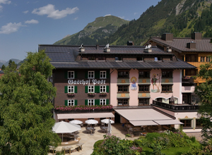 Ein alpenländliches Gasthaus mit dem Schriftzug „Gasthof Post“ ist abgebildet. Das Gebäude zeigt eine Mischung aus traditionellem und modernem Design, umgeben von Bäumen und mit einer Terrasse. Im Hintergrund sind grüne Berge sichtbar, was eine entspannte Atmosphäre vermittelt.