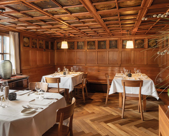 Ein gemütliches Restaurant mit Holzvertäfelung. Zwei Tische sind liebevoll mit weißen Tischdecken, Geschirr und Weingläsern gedeckt. An der Wand hängen Bilder, und die Beleuchtung ist warm und einladend. Der Boden besteht aus Holzparkett, was zur Atmosphäre beiträgt.