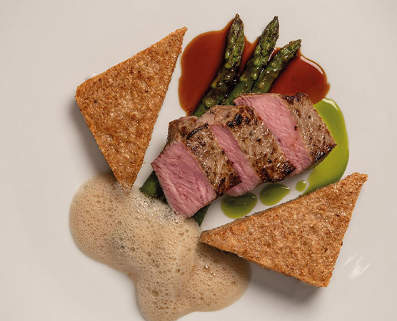 Ein Teller mit gegrilltem Fleisch, das in Scheiben geschnitten ist, liegt auf grünem Spargel. Daneben befinden sich drei Stücke knuspriges Brot und eine schäumende Sauce. Bunte Saucenränder ergänzen das Gericht.