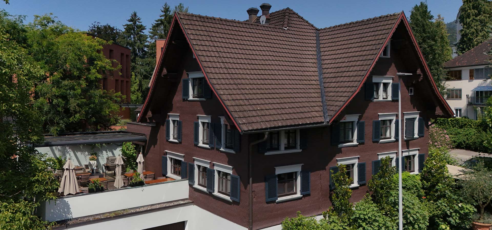 Ein braunes Haus mit einem steilen, roten Dach. Die Fassade hat mehrere Fenster mit weißen Rahmen und blauen Fensterläden. Im Vordergrund gibt es eine Terrasse mit Tischen, Stühlen und Sonnenschirmen. Um das Haus sind Bäume und Sträucher zu sehen, bei klarem, blauem Himmel.