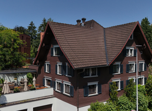 Ein mehrstöckiges, braunes Wohnhaus mit einem Satteldach ist sichtbar. Auf der linken Seite gibt es eine Terrasse mit Sonnenschirmen. Um das Haus sind Bäume und Sträucher zu sehen, und der Himmel ist klar und blau.