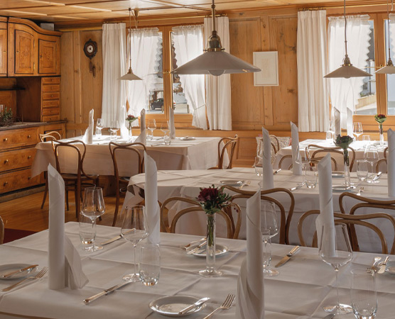 Ein gemütliches Restaurant mit Holzwänden. Tische sind mit weißen Tischdecken gedeckt, falteteServietten und Gläser stehen bereit. Die Fenster sind mit hellen Vorhängen dekoriert und die Atmosphäre ist einladend. Wasserflaschen und kleine Blumenarrangements sind auf den Tischen platziert.