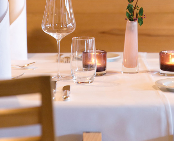 Ein elegant gedeckter Tisch in einem Restaurant, mit einem weißen Tischdecken. Darauf befinden sich ein Weinglas, ein Wasserglas, eine Kerze im Glas und eine kleine Vase mit grünen Zweigen. Besteck und Tellergeschirr sind sorgfältig arrangiert.