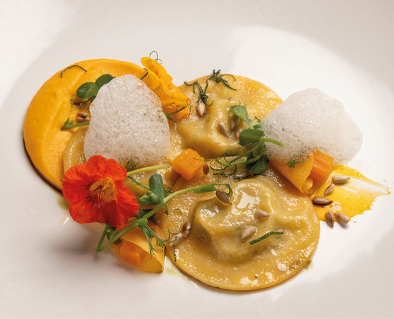 Ein elegantes Tellergericht mit Ravioli, umgeben von buntem Gemüse und grünen Kräutern. Auf der Oberfläche befinden sich leichte Schaumkugeln und geröstete Sonnenblumenkerne, die dem Gericht eine ansprechende Textur und Farbe verleihen.