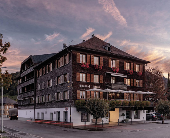 Das Bild zeigt ein mehrstöckiges Gebäude mit dem Namen "Gasthof Kene". Der Gasthof hat viele Fenster mit Holzläden und blühenden Pflanzen. Vor dem Gebäude sind einige Bäume und eine Straße zu sehen, unter einem farbenfrohen Abendhimmel.