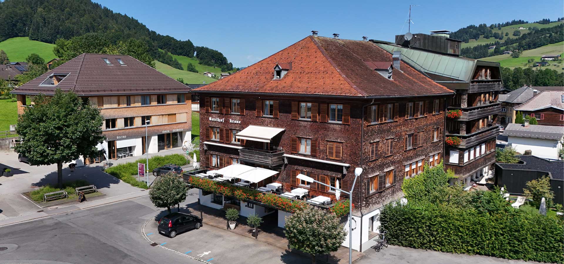 Ein traditionelles Gebäude mit einem rot-bräunlichen Holzfassade, das als Gasthof Krone dient. Vor dem Gebäude sind Tische und Stühle im Freien unter einem Markisenabdeckung. Die Umgebung zeigt grüne Wiesen und Hügel im Hintergrund.