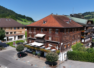 Ein traditionelles, mehrstöckiges Holzgebäude mit einem roten Satteldach, umgeben von Bäumen und gartengestalteten Flächen. Im Vordergrund sind Tische und Stühle auf einer Terrasse zu sehen. Es herrscht sonniges Wetter und die Umgebung ist ländlich.