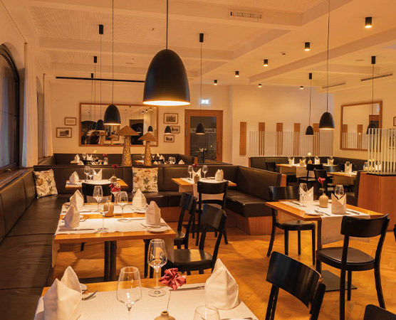 Ein stilvolles Restaurant mit Holztischen und bequemen Sitzbänken, elegant eingedeckt mit Gläsern und Servietten. Weiche Beleuchtung von modernen Deckenlampen schafft eine einladende Atmosphäre. An den Wänden hängen dekorative Bilder, die dem Raum einen geschmackvollen Touch verleihen.