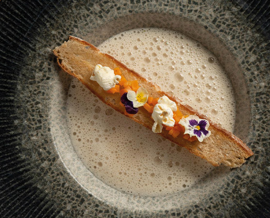 Ein längliches Stück Brot liegt auf einer dunklen Schale, belegt mit orangefarbenem Gemüse und garniert mit essbaren Blumen. Um das Brot schäumt eine helle, cremige Sauce, die den Teller umgibt.
