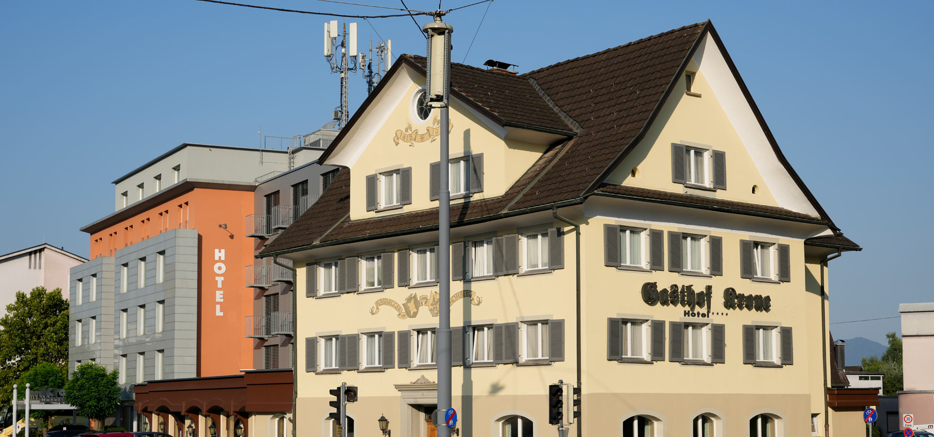 Eine Ansicht zweier Hotels im Stadtbild. Links steht ein älteres, gelb gestrichenes Gebäude mit vielen Fenstern und dem Namen "Gasthof Arne". Rechts ist ein moderner, grauer Bau mit der Aufschrift "HOTEL" in großen, weißen Buchstaben. Im Hintergrund ein blauer Himmel.
