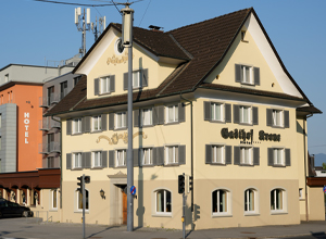 Das Bild zeigt ein zweigeschossiges, gelb gestrichenes Gebäude mit einem Satteldach und mehreren Fenstern. An der Fassade ist der Schriftzug "Gasthof Stern" deutlich sichtbar. Das Gebäude befindet sich an einer Straßenecke und wird von einer Straßenlampe und Verkehrsschildern umgeben.