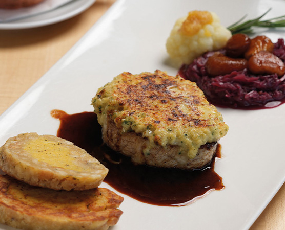 Ein Gericht auf einem langen weißen Teller, bestehend aus einem panierter Fleischstück, umgeben von einer dunklen Sauce. Neben dem Fleisch liegen zwei Scheiben einer gelben Beilage und darauf eine Portion Rotkohl mit Kastanien. Ein kleiner Blumenkohl-Türmchen ergänzt das Arrangement.