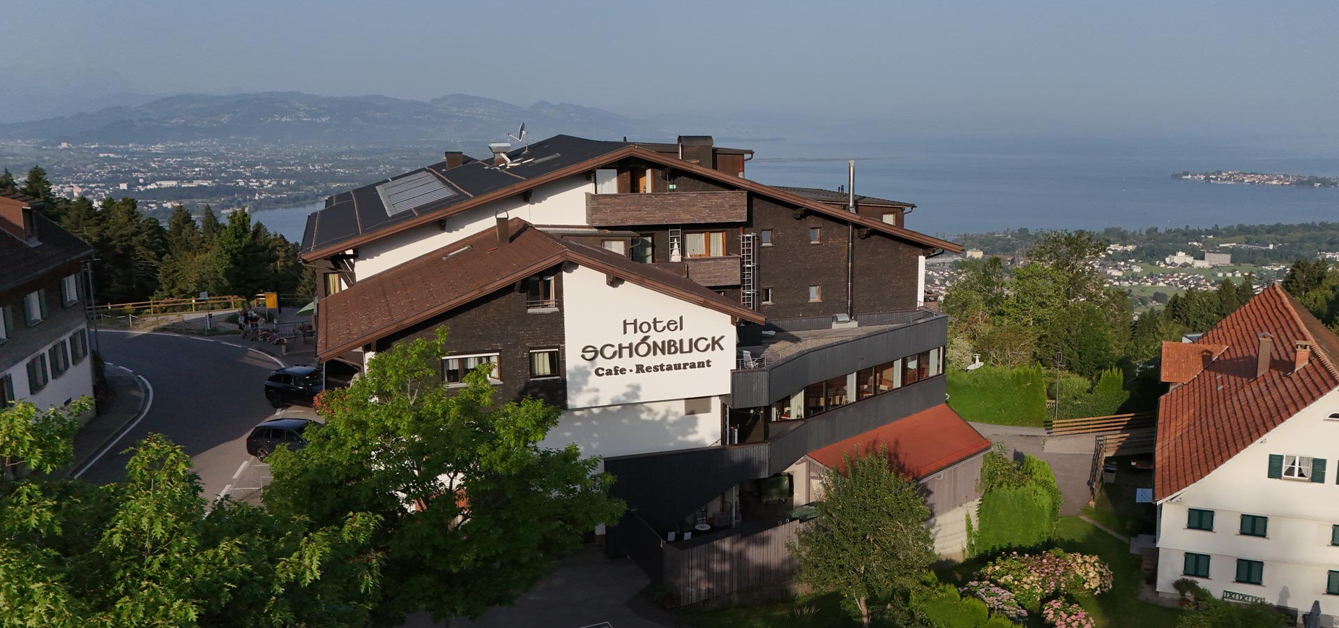 Das Bild zeigt das Hotel Schönblick, das über ein Café und Restaurant verfügt. Es befindet sich auf einem Hang mit Blick auf einen See und die umliegenden Berge. Die Architektur ist modern und umgeben von Bäumen und Grünflächen.
