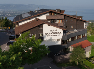 Das Bild zeigt das Hotel Schönbuck mit einem modernen, mehrstöckigen Gebäude. Der Schriftzug "Hotel Schönbuck Café - Restaurant" ist gut sichtbar. Im Vordergrund sind Bäume und ein Teil der Umgebung zu erkennen, während im Hintergrund ein Blick auf den Bodensee und die Landschaft zu sehen ist.