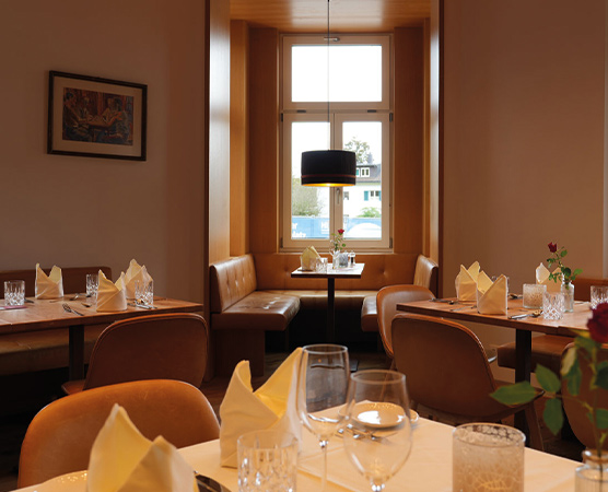 Ein gemütliches Restaurant mit Holzinterieur. Tische sind liebevoll gedeckt, mit Gläsern und Faltservietten. Im Hintergrund befindet sich ein rundes Fenster mit Blick nach draußen und eine moderne Lampe hängt von der Decke. Es strahlt eine einladende Atmosphäre aus.