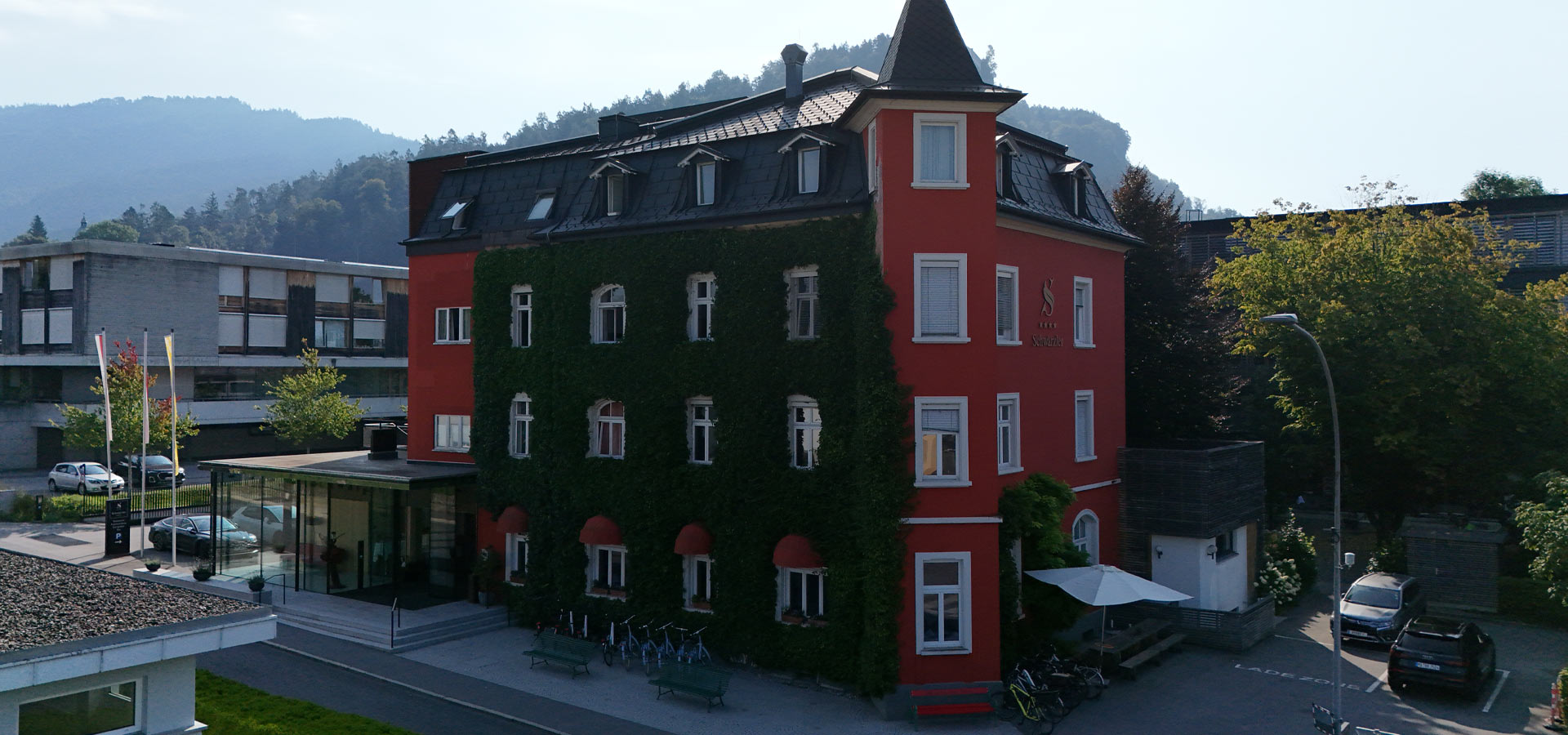 Ein rotes Gebäude mit mit Grün bewachsenen Wänden steht im Vordergrund. Im Hintergrund sind sanfte Hügel zu sehen. Vor dem Gebäude gibt es eine Treppe, Fahrradständer und einen Bereich mit kleinen Bäumen. Die Szene vermittelt eine ruhige, einladende Atmosphäre.