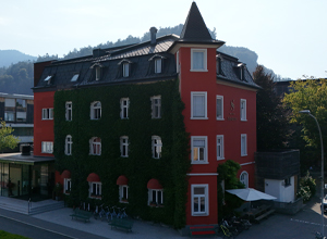Ein rotes Gebäude mit einem spitzen Dach, das teilweise von Grünpflanzen überwachsen ist. Es hat mehrere Fenster und Lichtstreifen. Im Vordergrund sind einige Sitzgelegenheiten und Fahrräder zu sehen. Die Umgebung ist von Bäumen und Hügeln geprägt.
