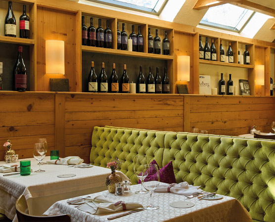 Ein elegantes Restaurant mit einer grünen, gepolsterten Sitzbank und einem Tisch gedeckt mit Gläsern und Servietten. Im Hintergrund stehen Weinflaschen in Regalen. Die Atmosphäre ist warm und einladend, mit sanftem Licht von Wandleuchten.
