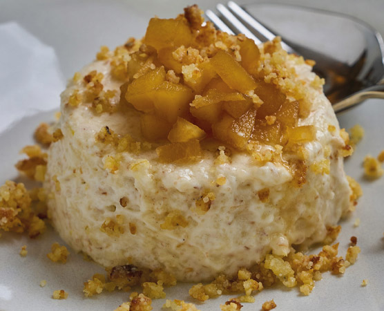 Ein cremiges Dessert mit einer Schicht aus fein gehackten, gebratenen Äpfeln und einer knusprigen Kruste aus Keksbröseln. Das Gericht ist leicht und süß, angerichtet auf einem weißen Teller mit einem Silberbesteck im Hintergrund.