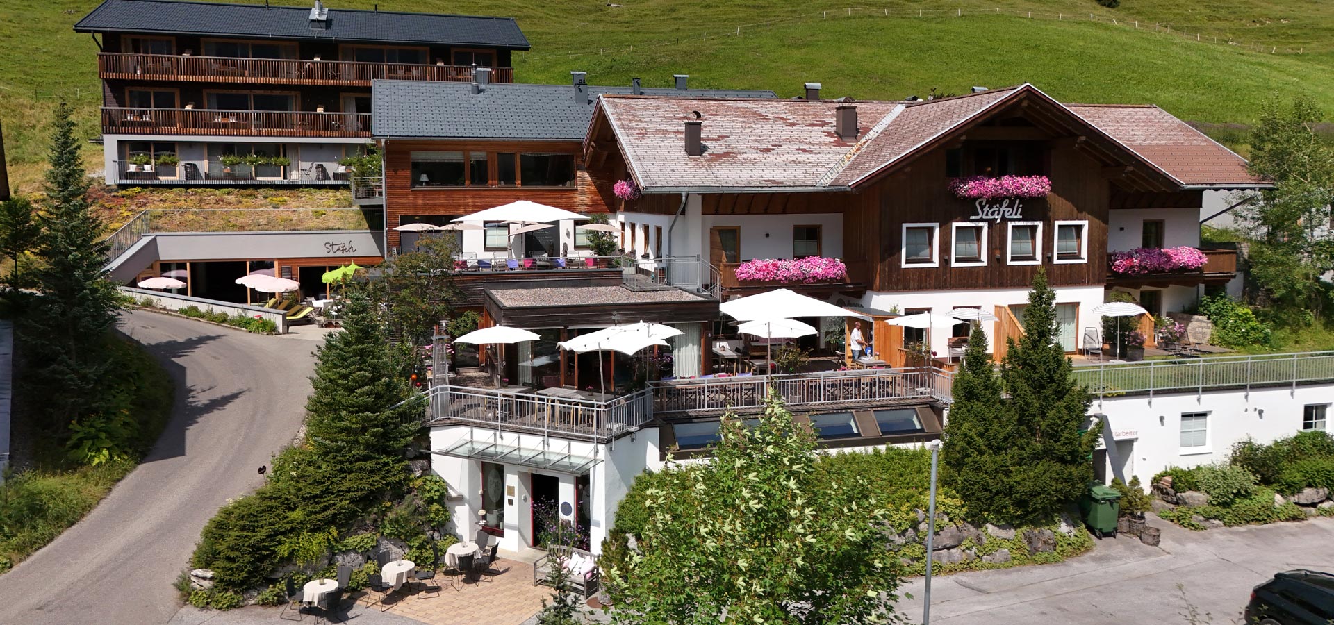 Das Bild zeigt ein rustikales Hotel mit mehreren Etagen, umgeben von grünen Wiesen. Auf dem Balkon und der Terrasse stehen Tische mit Sonnenschirmen. An der Fassade hängen Blumenkästen. Eine Straße führt zum Eingang des Hotels, der gut sichtbar ist.