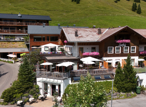 Das Bild zeigt ein alpines Hotel mit mehreren Gebäuden. Die Fassaden sind aus Holz und die Balkone sind mit Blumen dekoriert. Vor dem Hotel sind Terrassen mit Sonnenschirmen und Tischen, umgeben von Bäumen und Wiesen. Ein malerischer Bergblick ist im Hintergrund sichtbar.