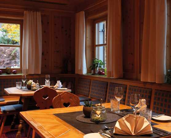 Der Innenraum eines gemütlichen Restaurants mit Holzwänden. Ein Tisch ist festlich gedeckt mit Gläsern, Besteck und kunstvoll gefalteten Servietten. Im Hintergrund sind Pflanzen und Fenster mit Vorhängen sichtbar. Das Ambiente wirkt warm und einladend.