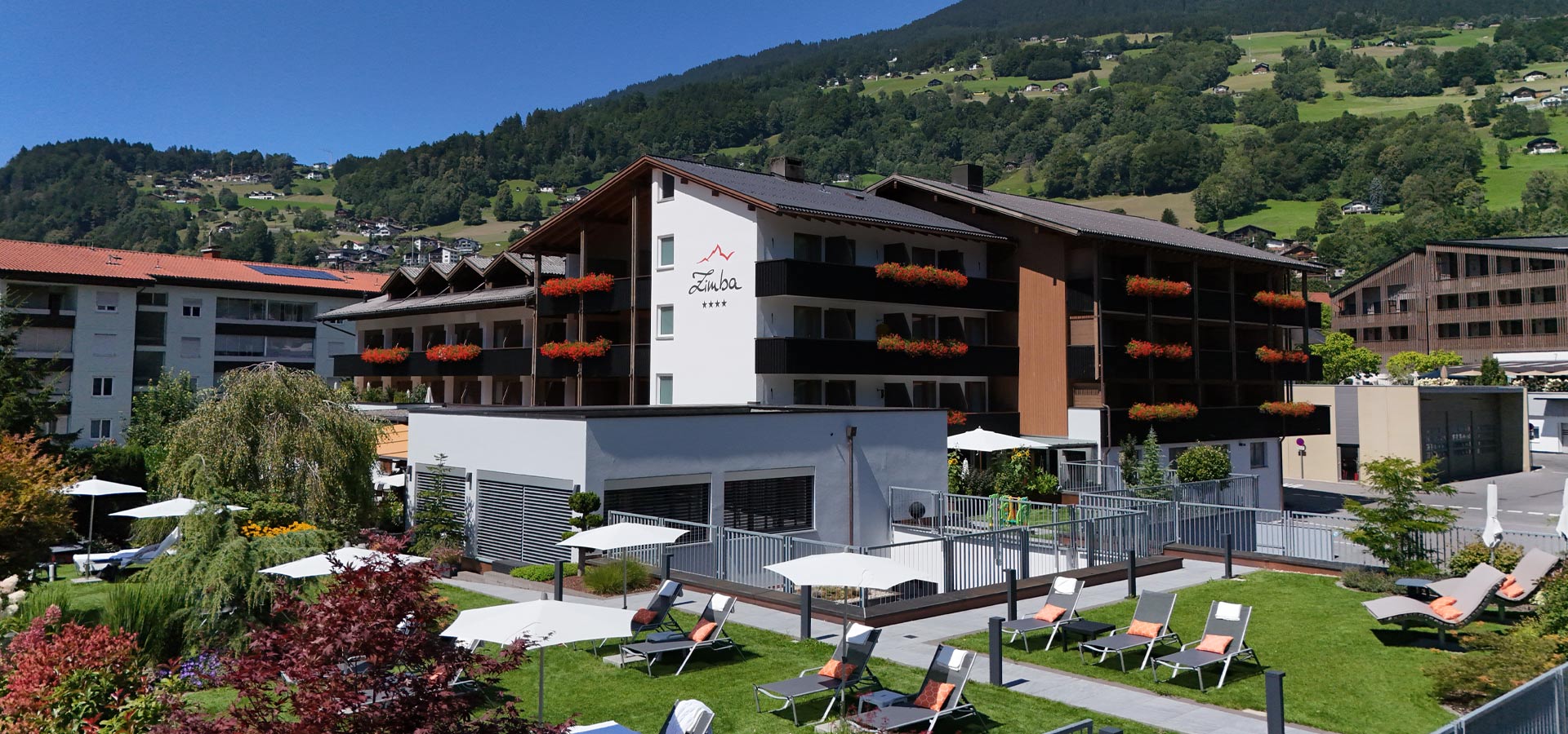 Das Hotel Zimba liegt in einer malerischen Umgebung, umgeben von Bergen und Grünflächen. Die Fassade des Hotels ist modern mit Blumenkästen. Im Vordergrund befinden sich Liegen auf einem gepflegten Rasen und Sonnenschirme, die eine einladende Atmosphäre schaffen.