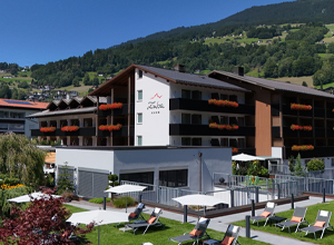 Ein modernes Hotelgebäude mit mehreren Stockwerken, umgeben von einer grünen Landschaft und Bergen im Hintergrund. Der Eingangsbereich ist freundlich gestaltet, und auf der Terrasse stehen Liegen zur Verfügung. Blühende Pflanzen hängen von den Balkonen.