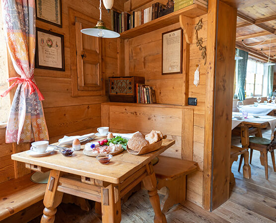 Gemütliche hölzerne Bauernstube mit kleinem Tisch, gedeckt für Frühstück: Brot, Eier, Tassen, Marmelade und Blumen; rustikale Holzwände und Sitzbank.
