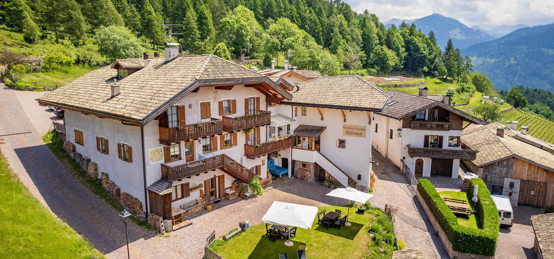 Alpines Gästehaus-Ensemble mit weiß verputzten Gebäuden und Holzbalustraden, Innenhof mit Sitzplätzen und Sonnendach, eingebettet in grüne Wiesen, Nadelwald und dahinter aufragende Berggipfel.