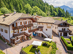 Luftaufnahme eines traditionellen Alpenhotels mit weißen Wänden und Holzbalkonen, zentraler Innenhof mit Rasen, Tischen, Sonnenschirm und Liegestühlen, eingebettet in grüne Wiesen und bewaldete Berge.