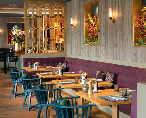 Ein elegantes Restaurant mit Holzwänden und einer lila gepolsterten Bank. Tische sind ordentlich mit Geschirr und Dekorationen eingedeckt. An den Wänden hängen Bilder von Pflanzen, und dekorative Blumenarrangements stehen in Vasen. Sanfte Beleuchtung schafft eine einladende Atmosphäre.