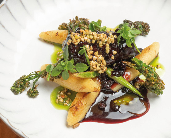 Ein Teller mit gebratenen, goldbraunen Stücken, garniert mit einer fruchtigen Sauce aus dunklen Beeren. Dazu sind grüne Gemüsestreifen und knuspere Kräuter dekoriert, die dem Gericht eine frische Note verleihen. Die Anordnung ist ansprechend und farbenfroh.