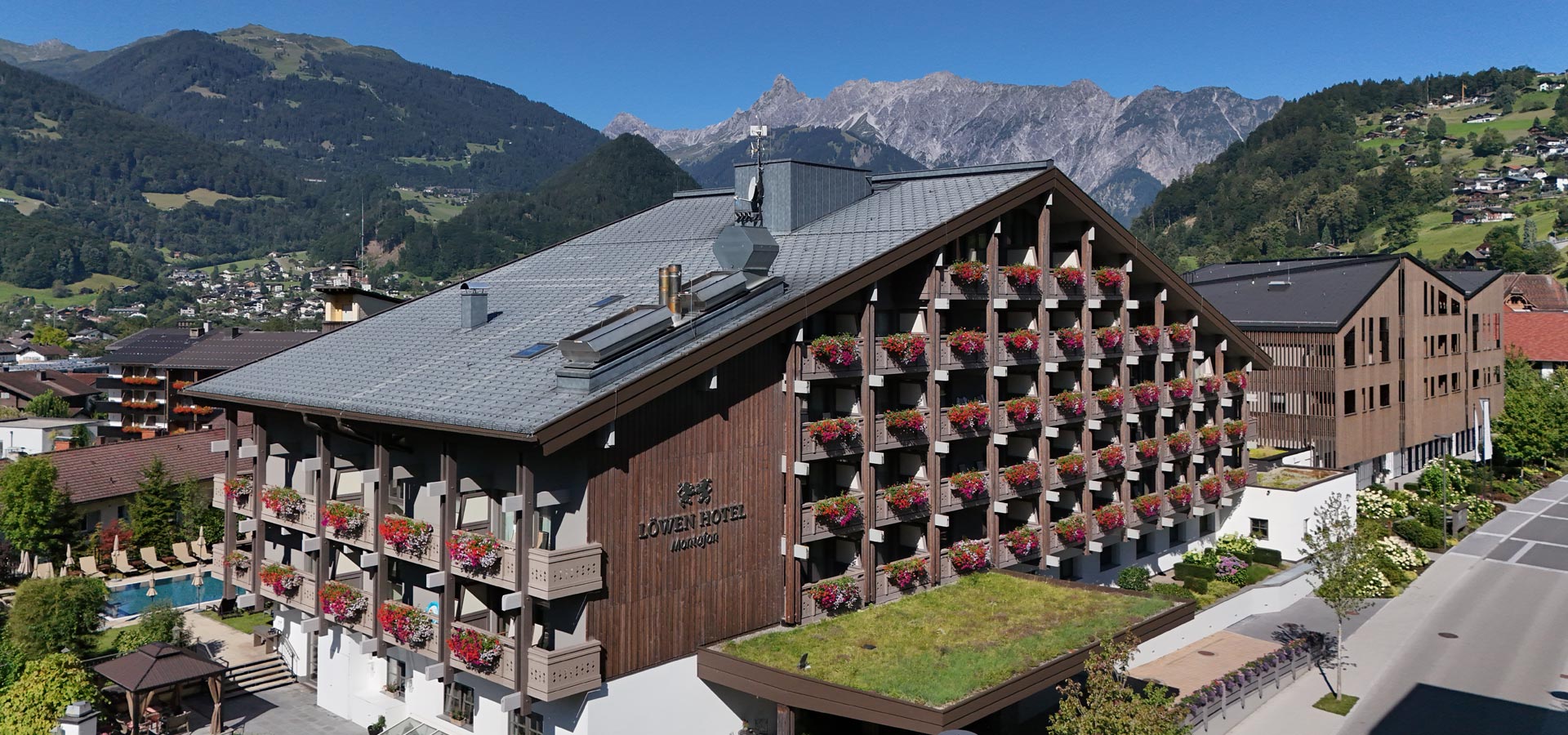 Das Bild zeigt das Löwen Hotel in Mandly, ein modernes Holzgebäude mit Blumenkästen an den Balkonen. Im Hintergrund sind majestätische Berge und ein klarer blauer Himmel zu sehen. Der Bereich vor dem Hotel ist bepflanzt und umfasst einen Pool.