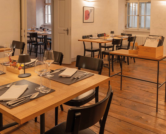 Ein gemütliches Restaurant mit Holztischen und schwarzen Stühlen. Auf einem Tisch sind Gläser, Besteck und Servietten gedeckt. Im Hintergrund sind weitere Tische sichtbar, und an der Wand hängt ein Kunstwerk. Der Boden ist aus Holz und die Wände sind hell gestrichen.