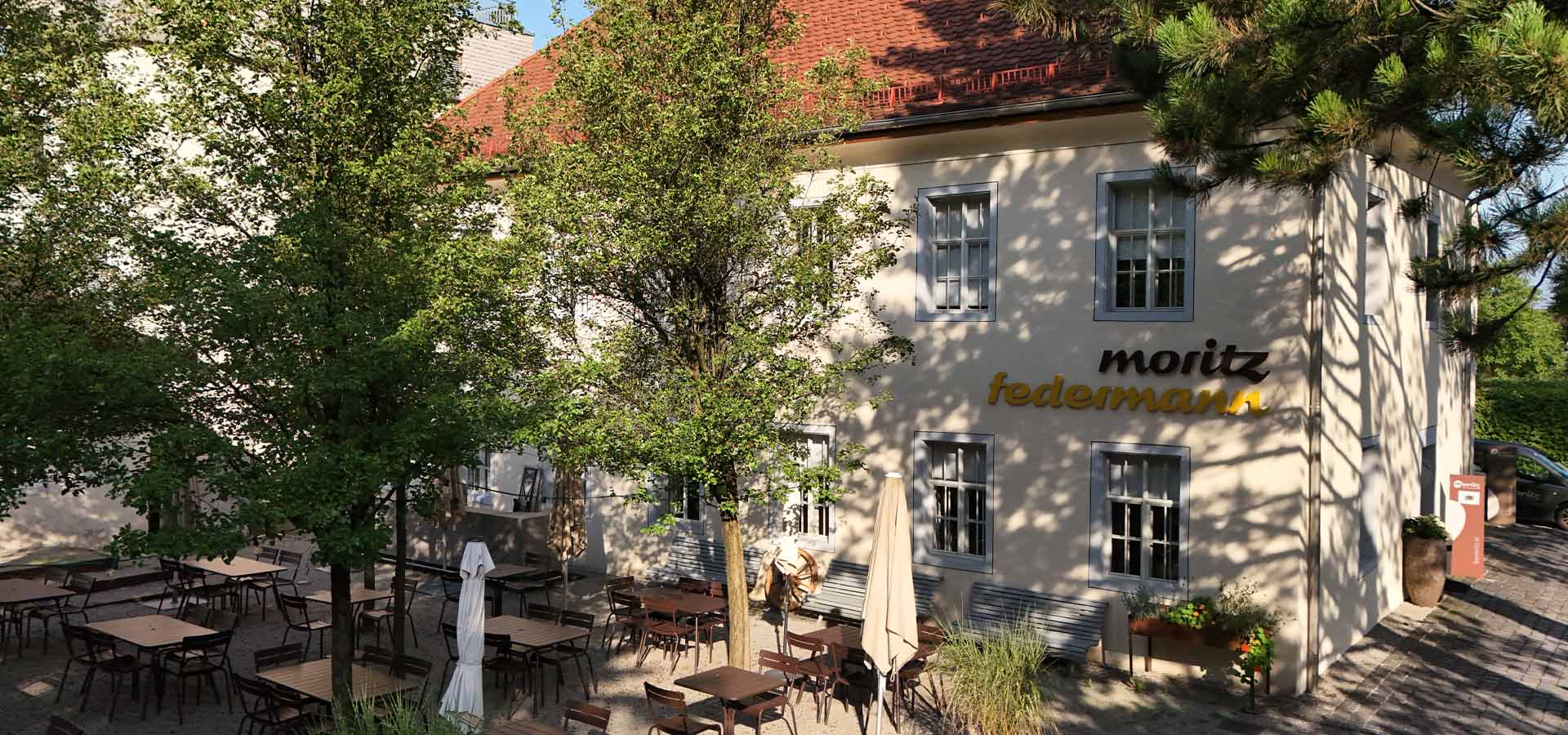 Das Bild zeigt ein Restaurant mit Außenbereich, umgeben von Bäumen. Mehrere Tische und Stühle sind im Freien aufgestellt, einige Sonnenschirme sind geschlossen. An der Wand des Restaurants steht der Name "Moritz Federmann" in Verbindung mit hellem Licht.