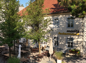 Das Bild zeigt ein Restaurantgebäude mit einem roten Dach. Vor dem Gebäude befinden sich zahlreiche Tische und Stühle auf einem Außenterrassenbereich. Über dem Eingang steht der Schriftzug „moritz federman“. Bäume umgeben den Bereich und fügen eine grüne Note hinzu.